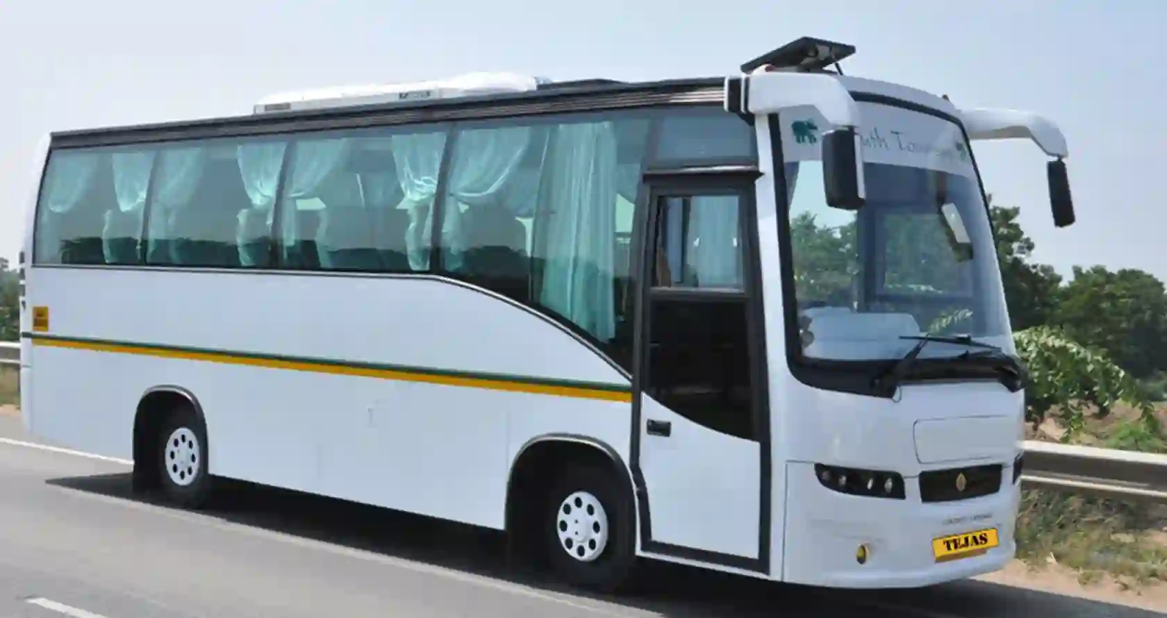 A white tour bus with large windows and curtains parked by the roadside, surrounded by greenery under a clear sky.