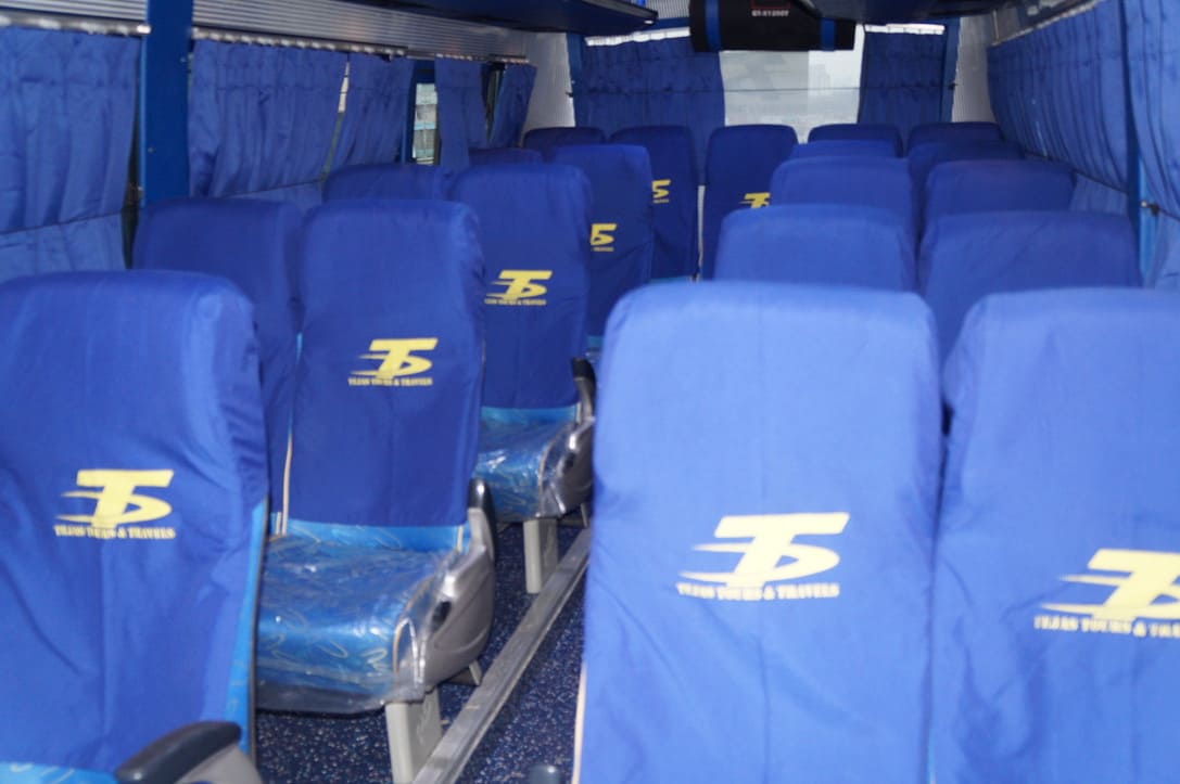 Empty interior of a bus, featuring rows of blue seats with logos, curtains on the windows, and a mounted screen above.
