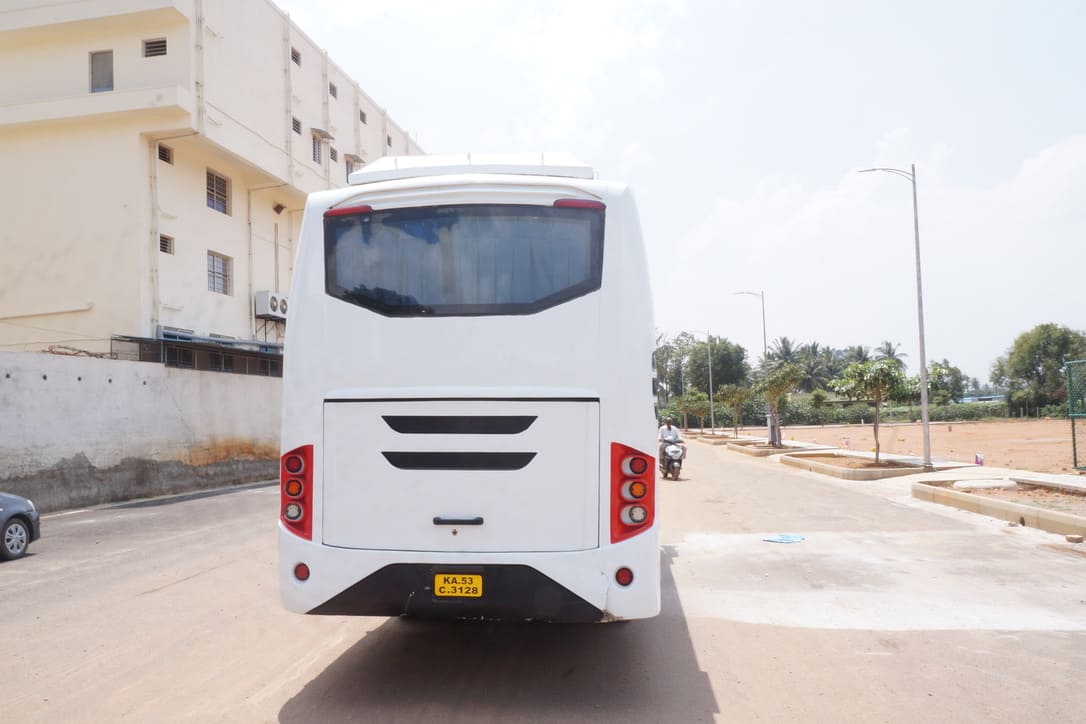 A white bus viewed from the rear, parked on a road beside a building, with trees and a motorcycle in the background.
