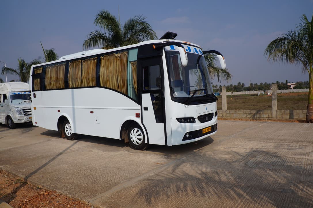 A white travel bus with curtains is parked on a paved area, surrounded by palm trees and open land under a clear sky.