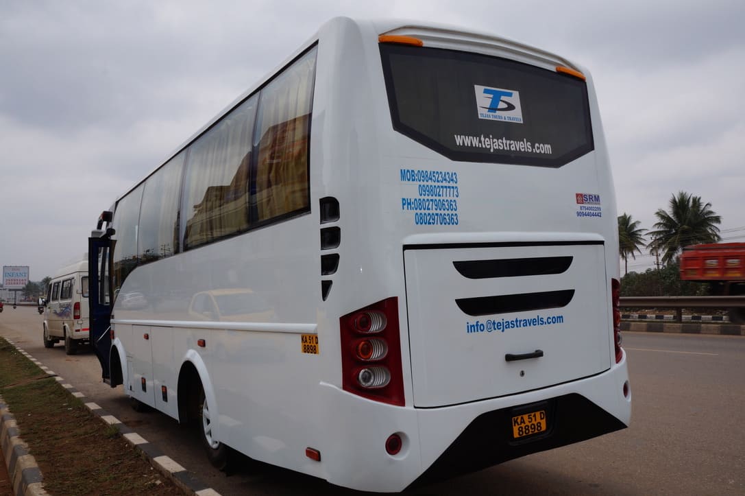 A white bus parked on a roadside, displaying travel service contact details, with palm trees and vehicles in the background.