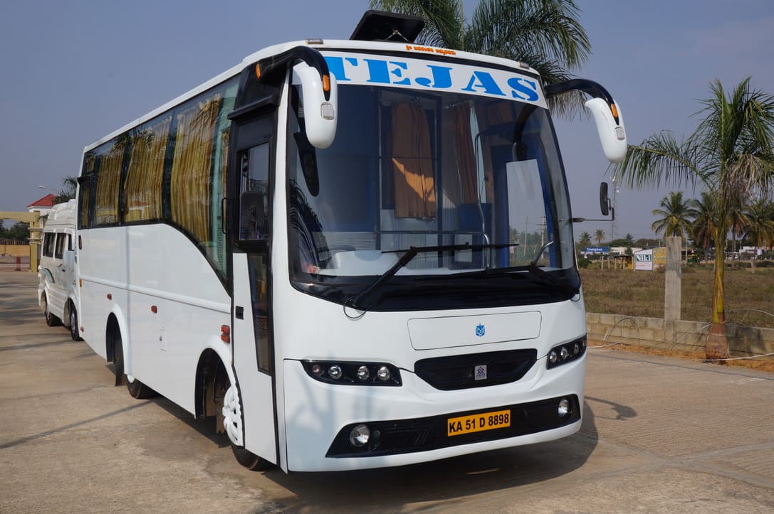 A white bus with the name "TEJAS" on the front, parked on a concrete surface beside palm trees and a grassy area.