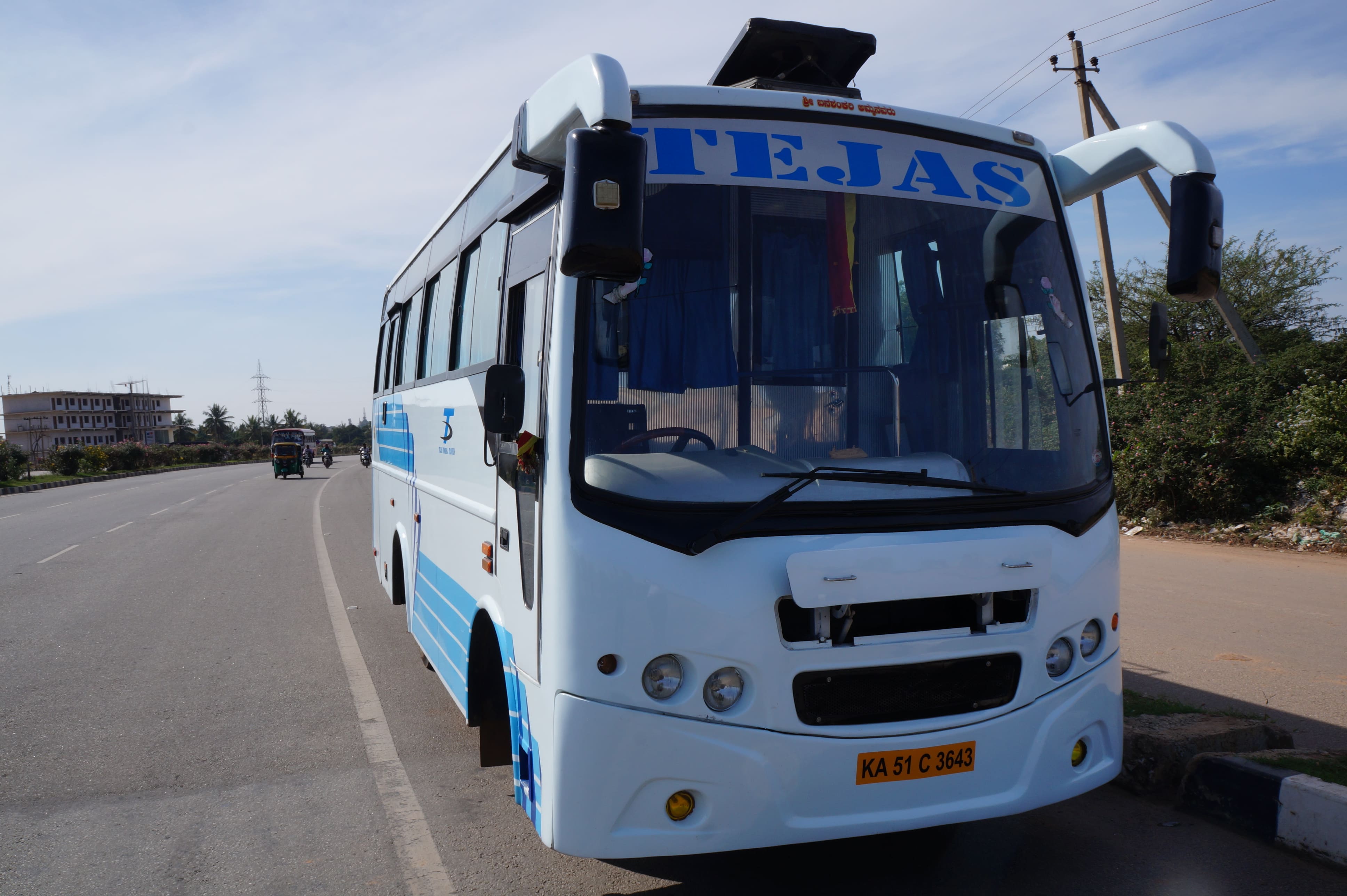 A blue and white bus named "Tejas" parked beside a roadside with buildings and trees in the background under a clear sky.