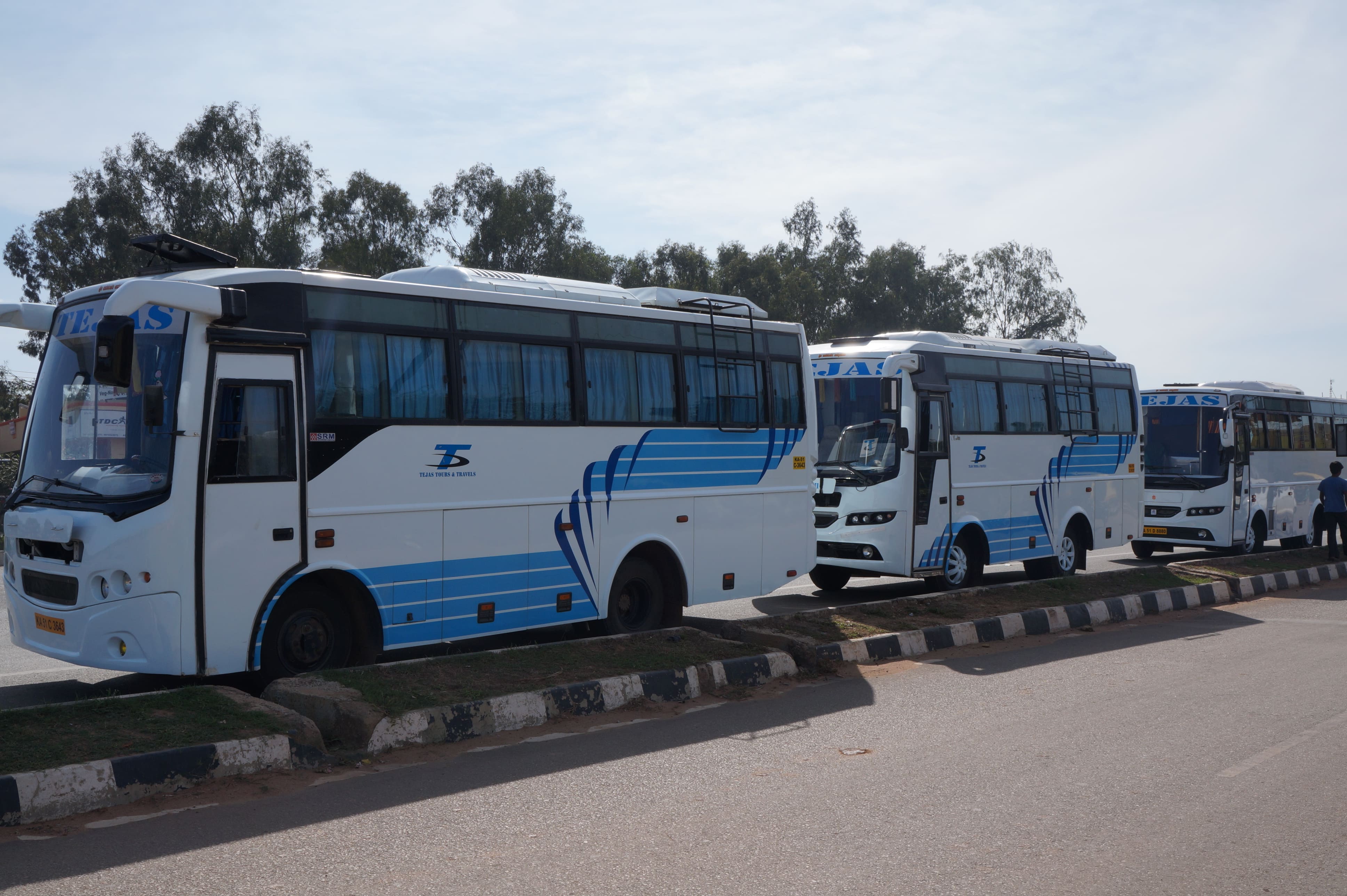 Three white buses with blue accents parked along a roadside, surrounded by trees and a clear sky.