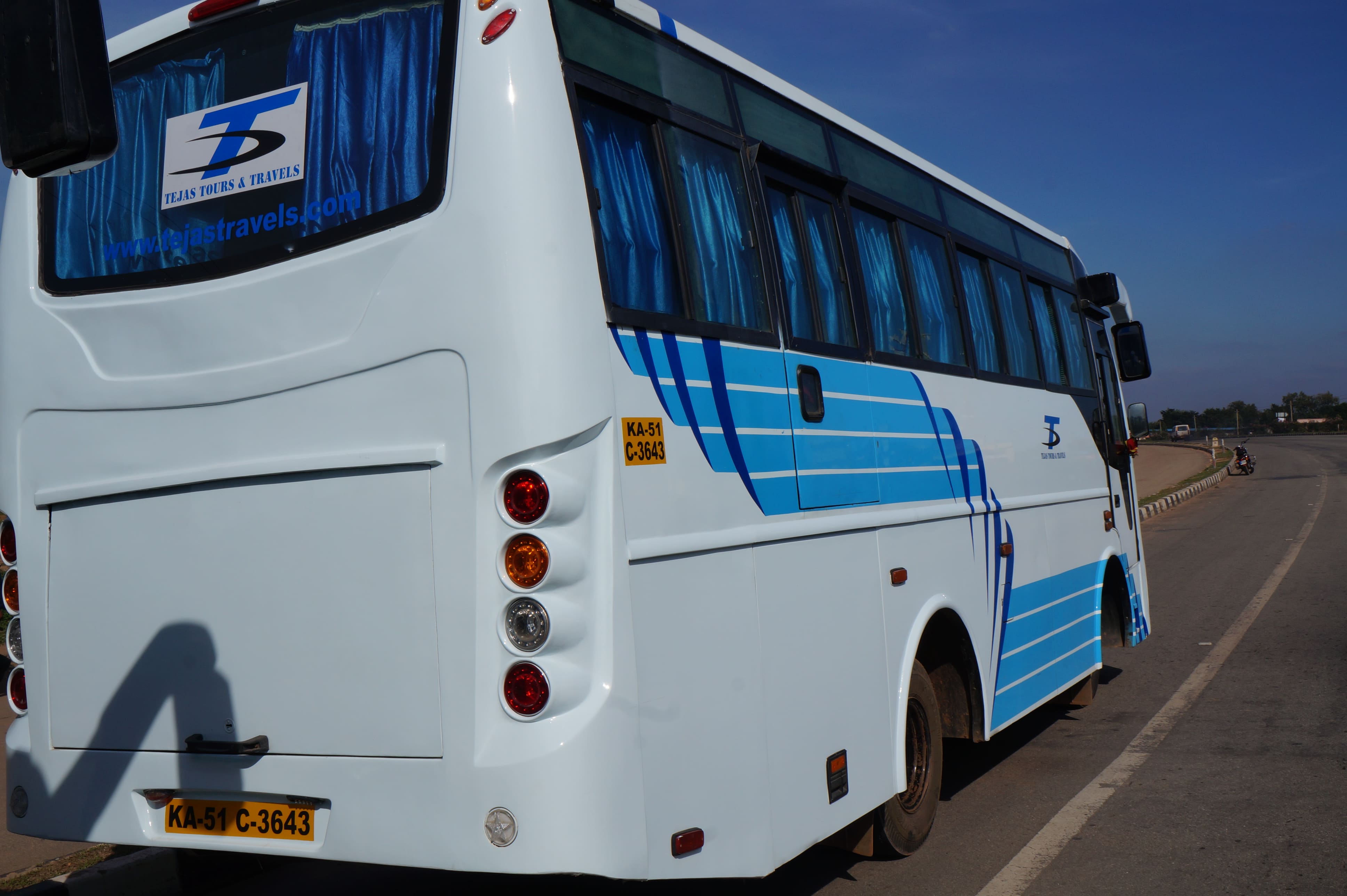 White travel bus with blue stripes parked by the roadside, displaying "Tejas Tours & Travels" on the rear window.