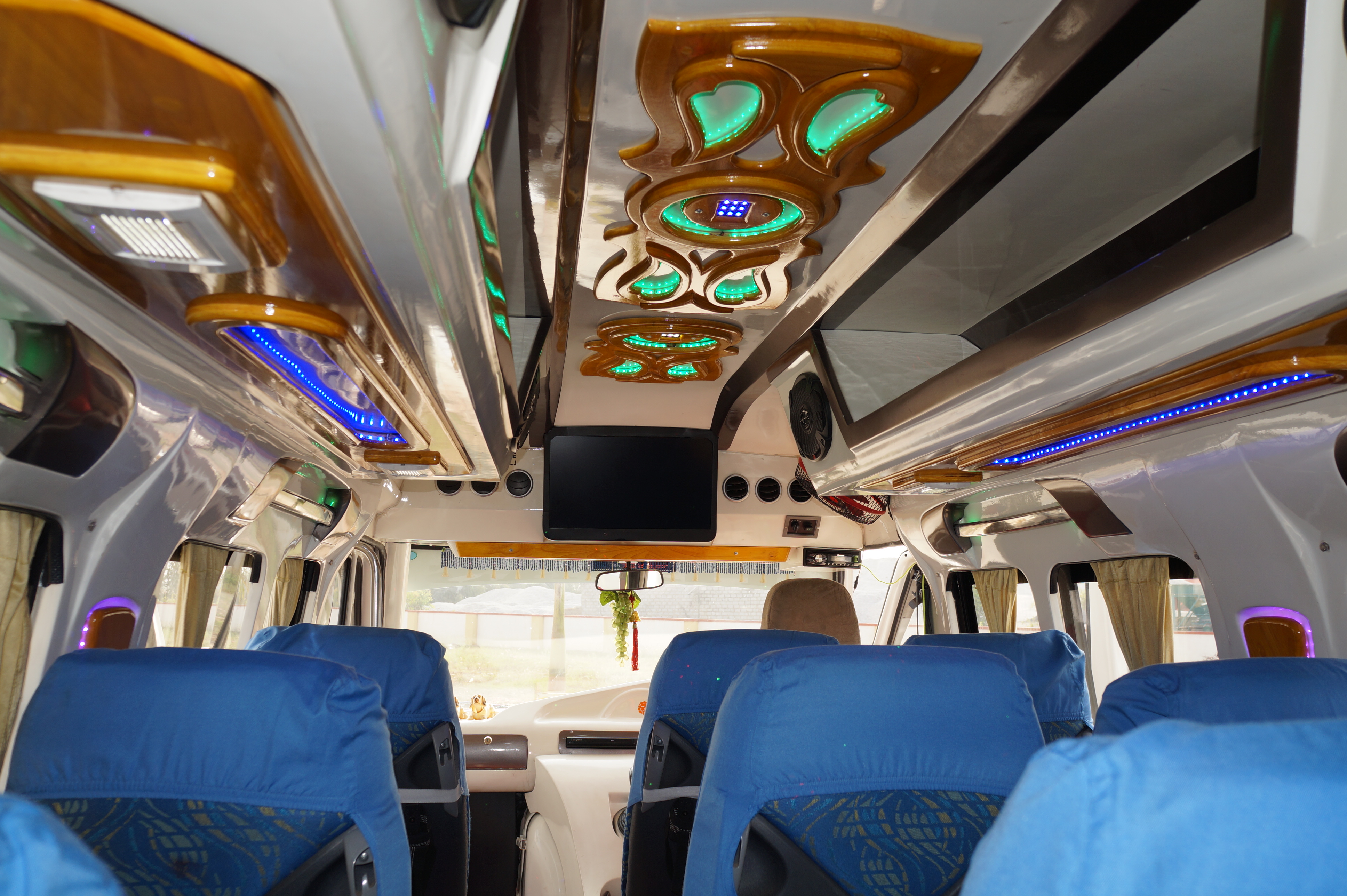 Interior of a minibus featuring blue seat covers, decorative lighting, a ceiling design, and a screen at the front.