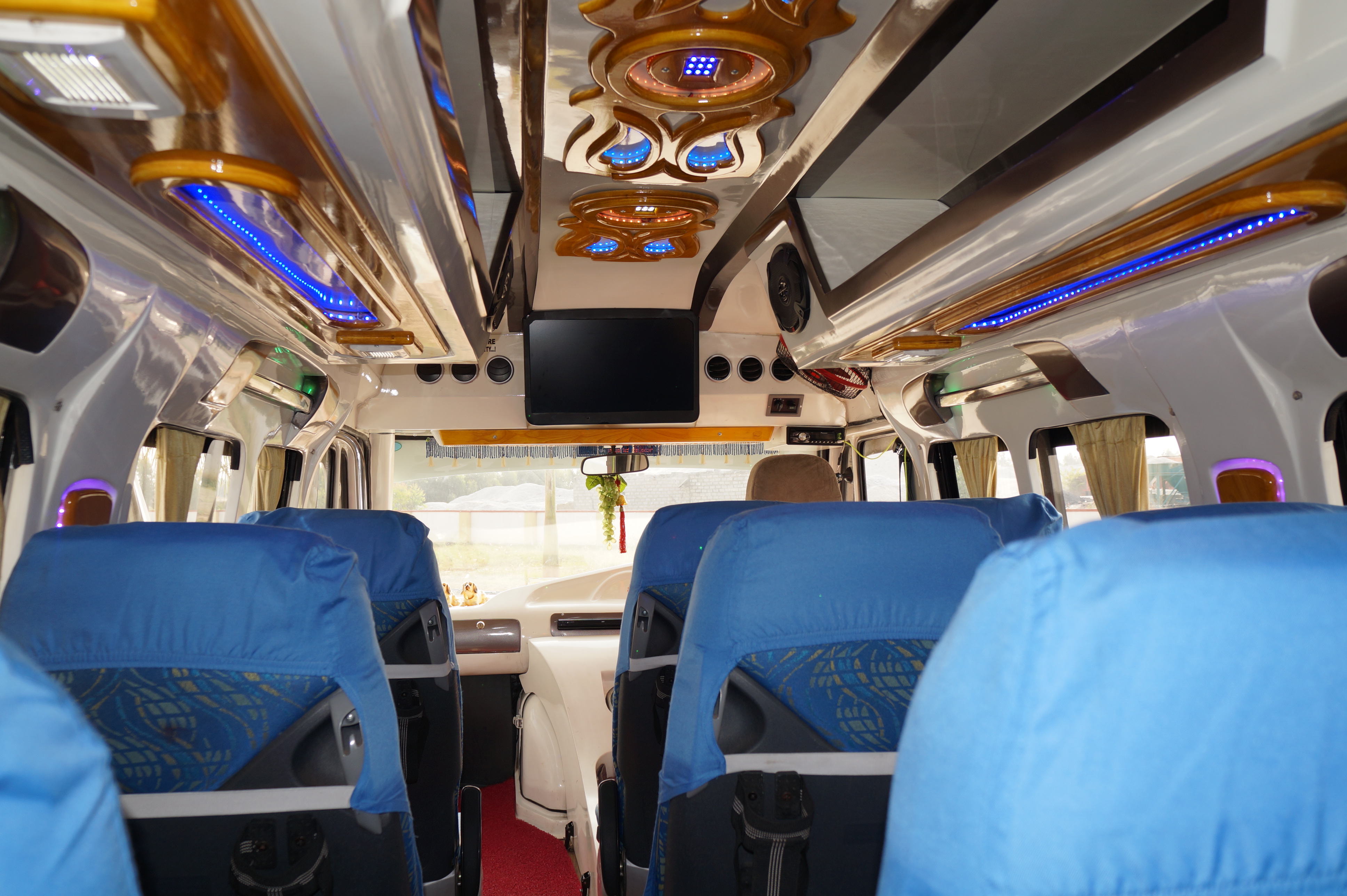 Interior of a spacious, brightly lit vehicle with blue seats, wooden accents, and a mounted screen at the front.