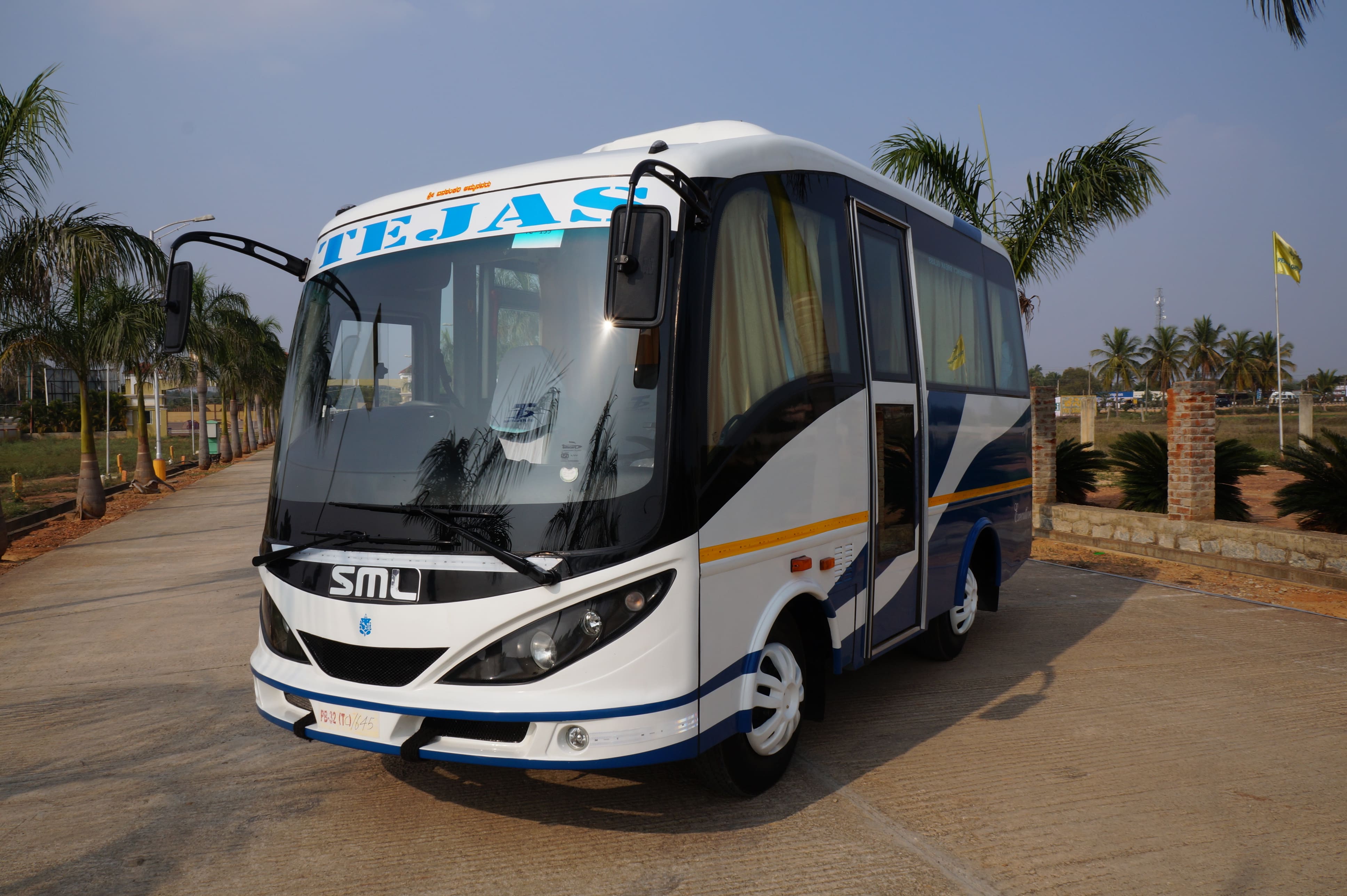 A white and blue bus with "TEJAS" written on it parked on a concrete path lined with palm trees and greenery.