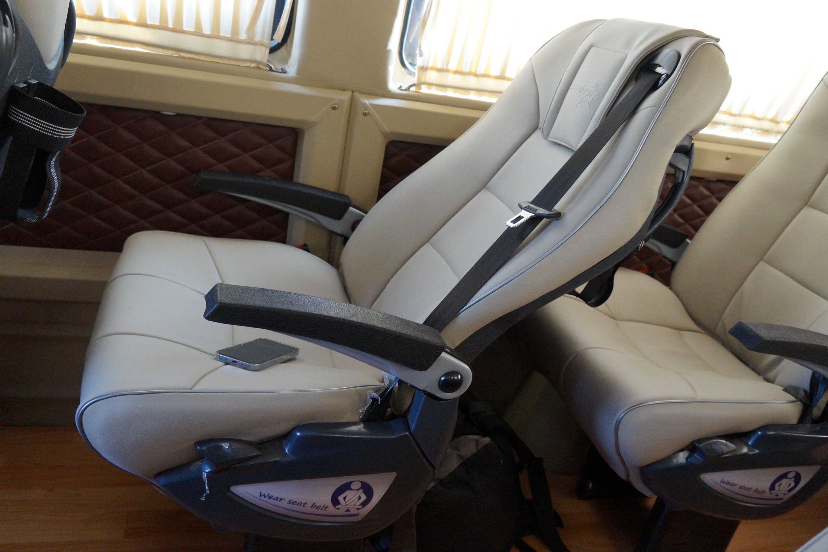 A modern, beige airplane seat with a seatbelt visible, next to a dark bag and a smartphone resting on the armrest.