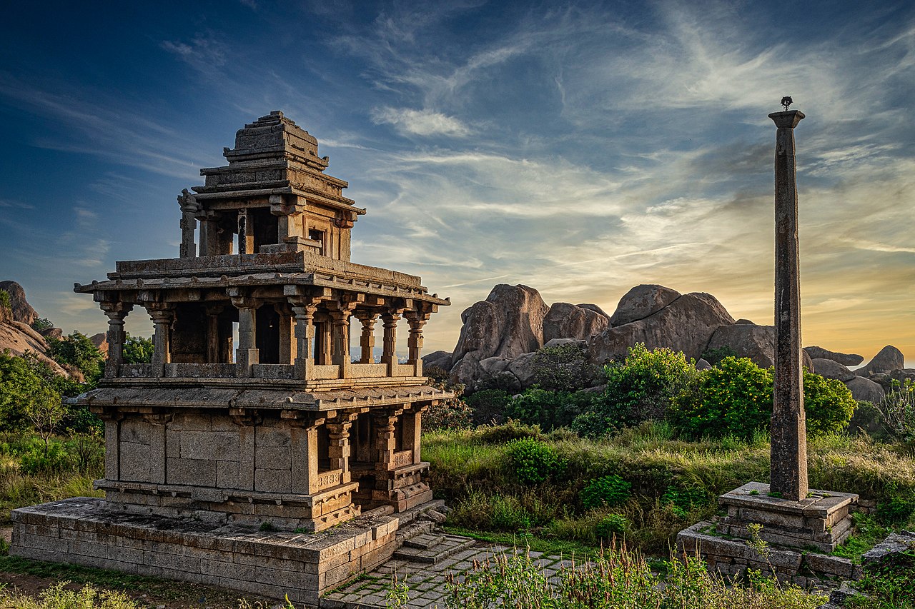 Ancient stone temple with intricate pillars, beside a tall column, set against rocky hills and a vibrant sunset sky.