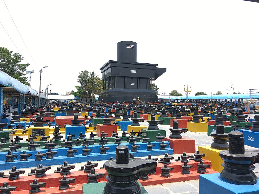 A temple complex featuring numerous colorful Shiva lingams arranged in rows, with a large black structure in the background.