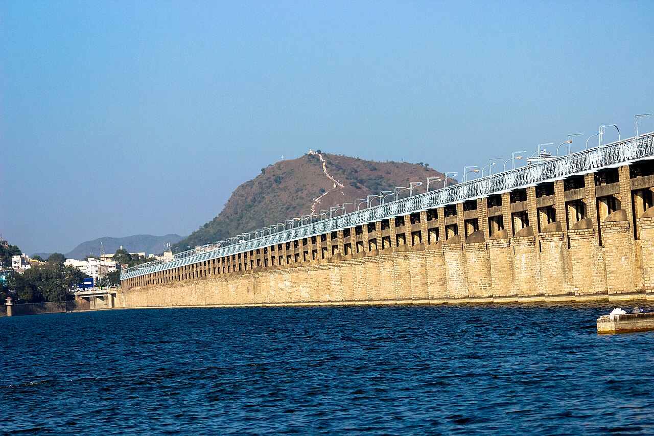 A long stone dam stretches across a blue lake, with a hillside and path visible in the background under a clear sky.
