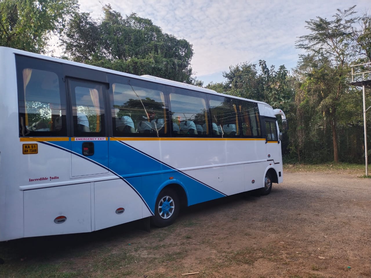 A white and blue bus is parked on a gravel surface, surrounded by green trees and shrubbery under a cloudy sky.