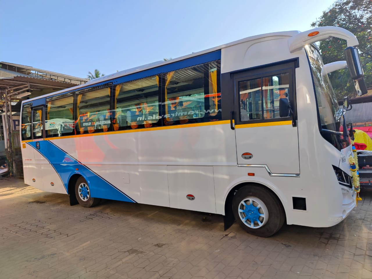 A modern white and blue bus parked at a depot, showcasing large windows and bright blue wheels under clear skies.