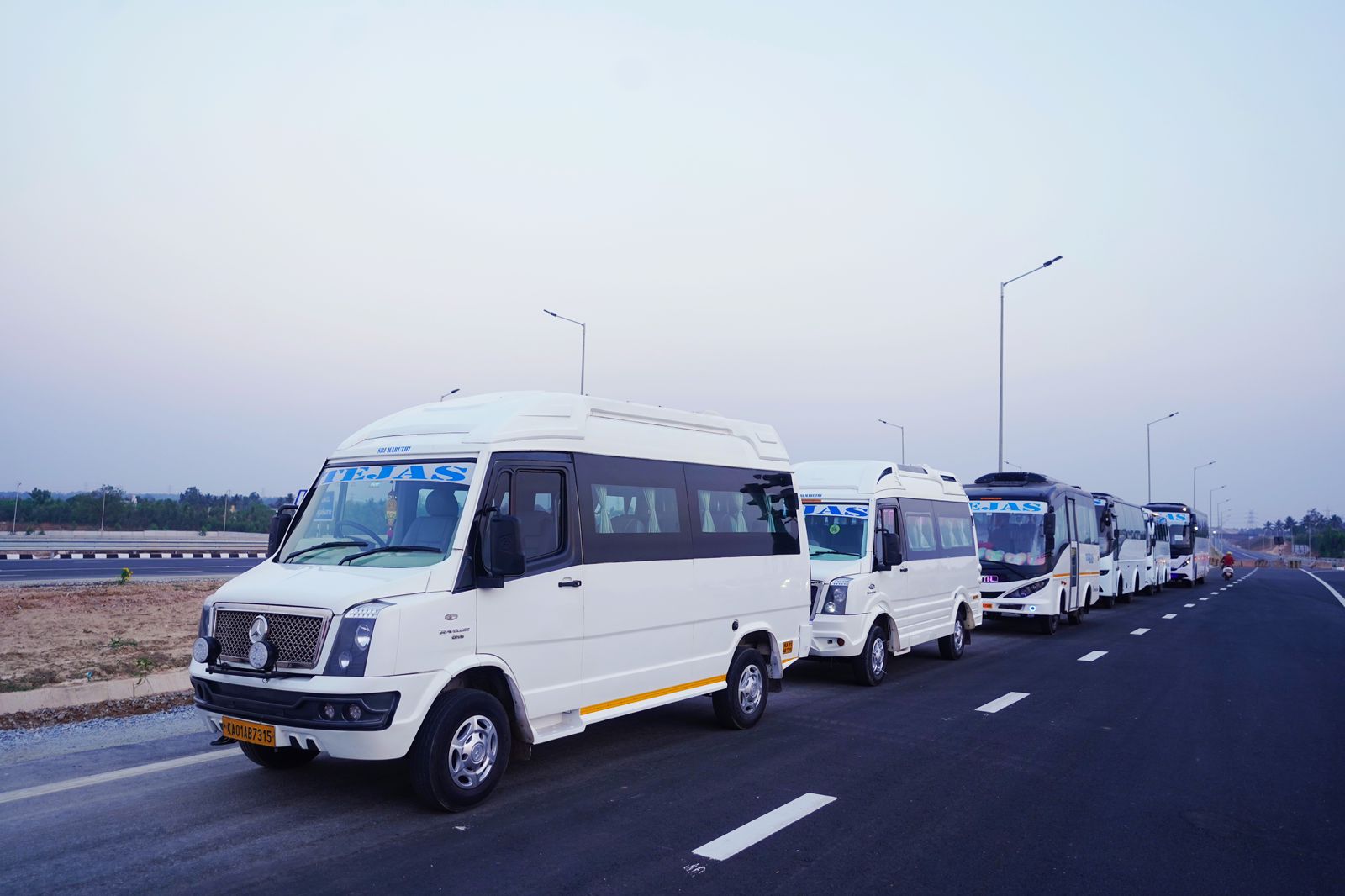 Six white vans and buses parked on a highway under a clear sky, with streetlights along the road.