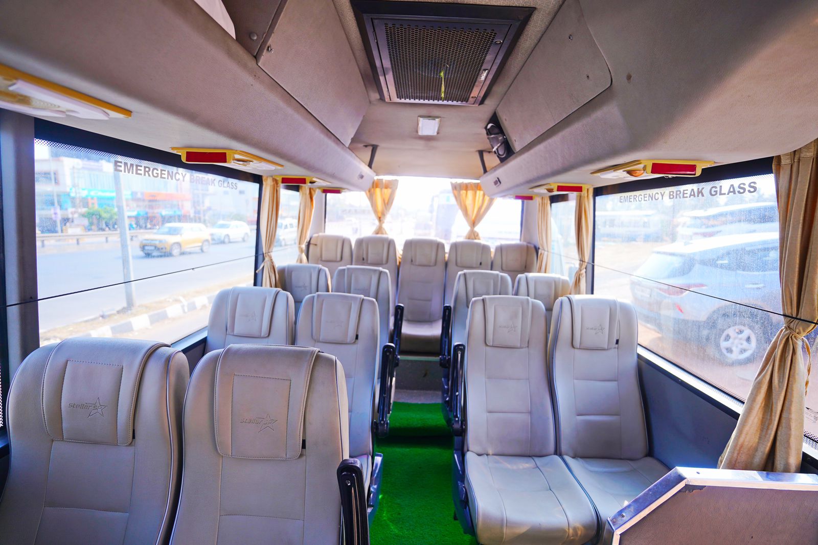 Interior of a spacious, empty bus with beige seats, green carpet, and sunlight streaming through the curtains.