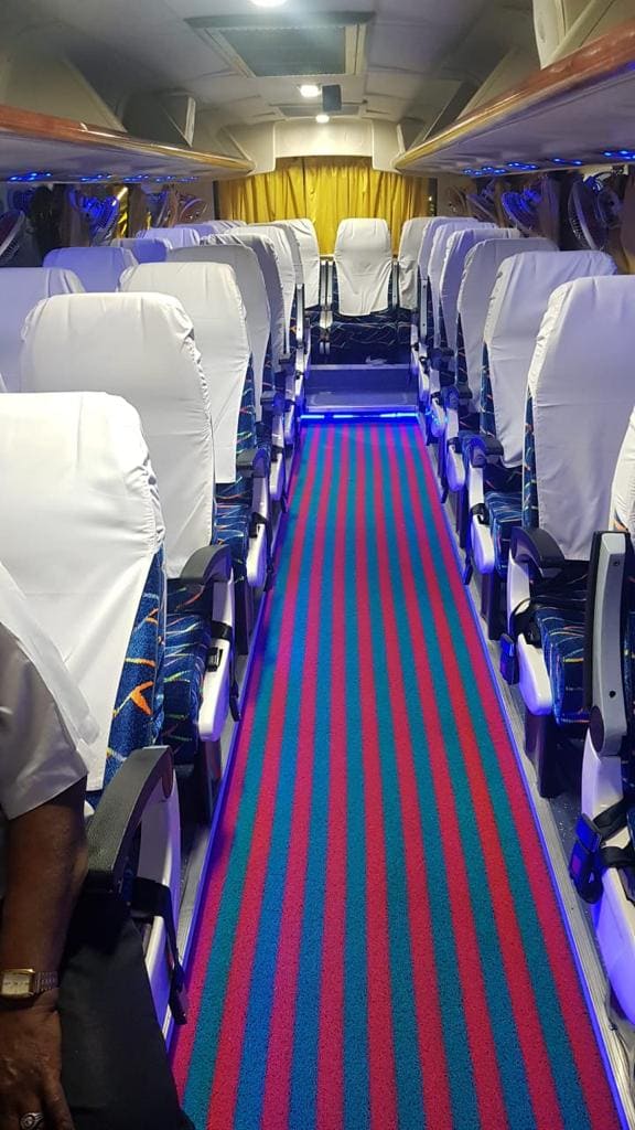 Interior of a bus featuring rows of white-covered seats, vibrant striped carpet, and blue ambient lighting.