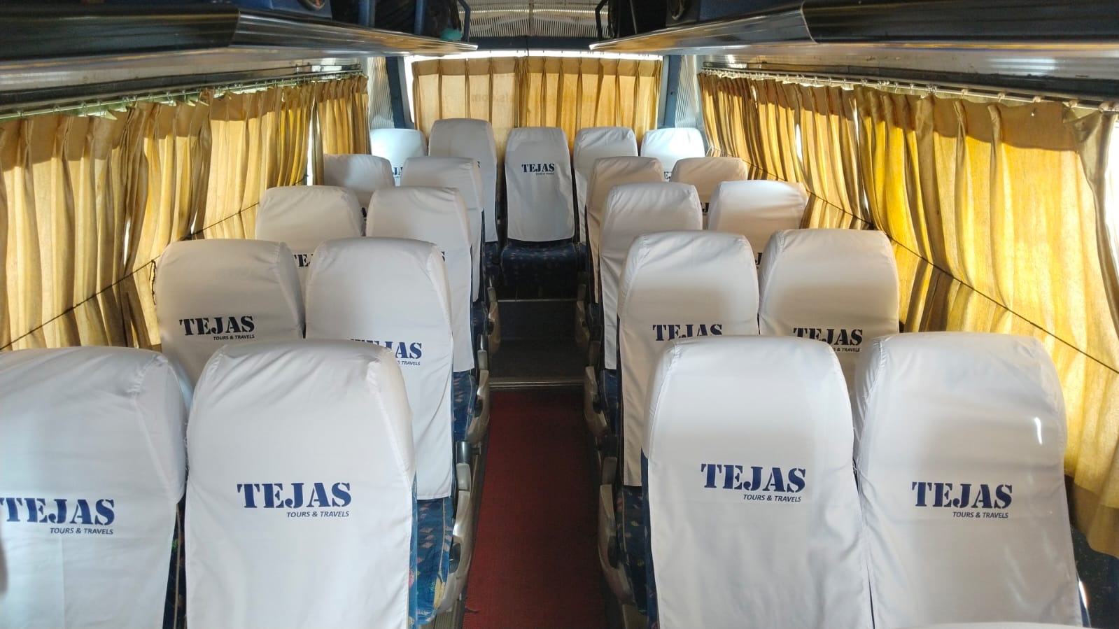 Interior of a bus featuring rows of white seats with "TEJAS" branding, warm lighting, and curtained windows.