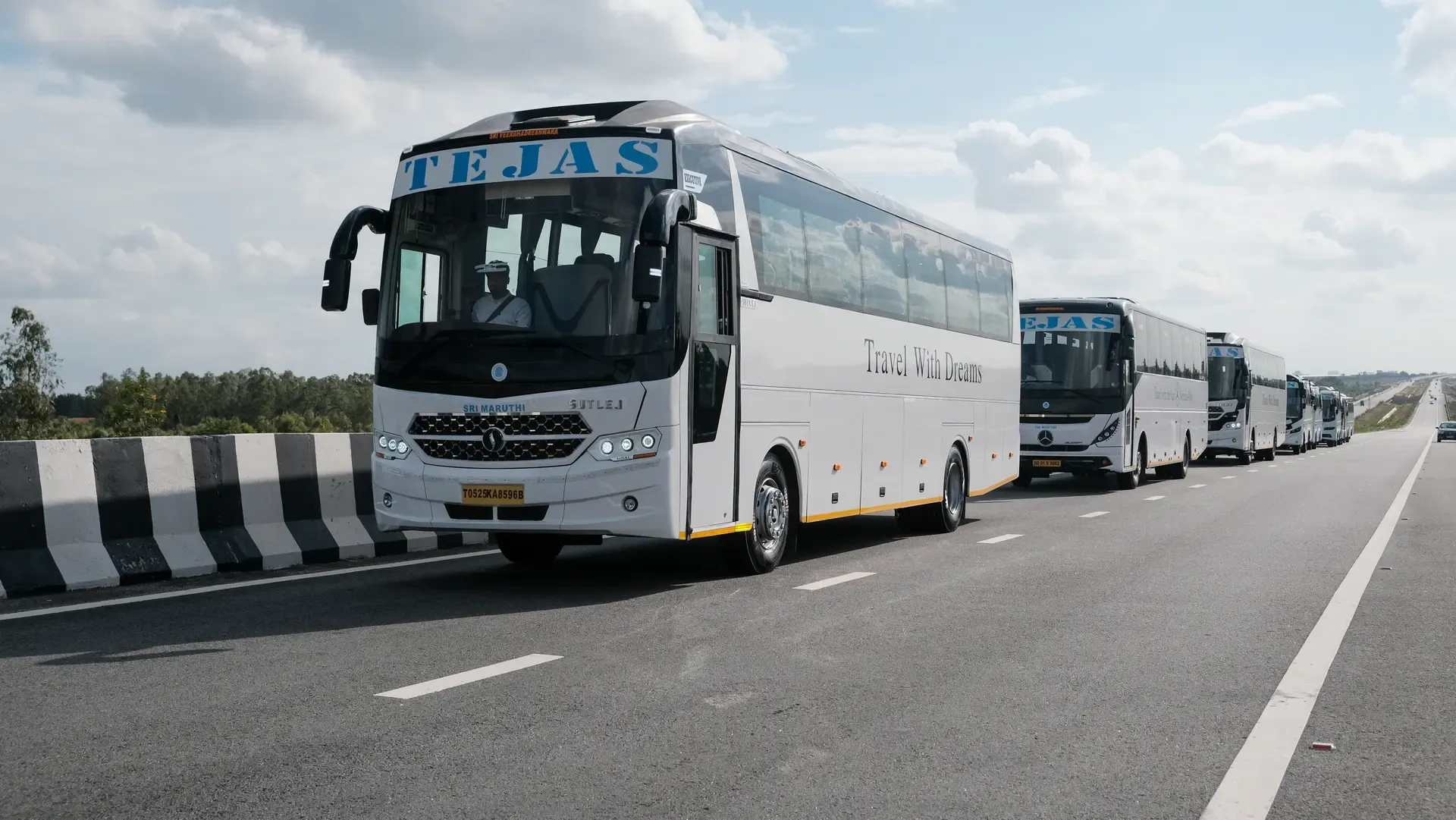 A fleet of white buses lined up on a highway under a cloudy sky, showcasing "TEJAS" branding on the front.