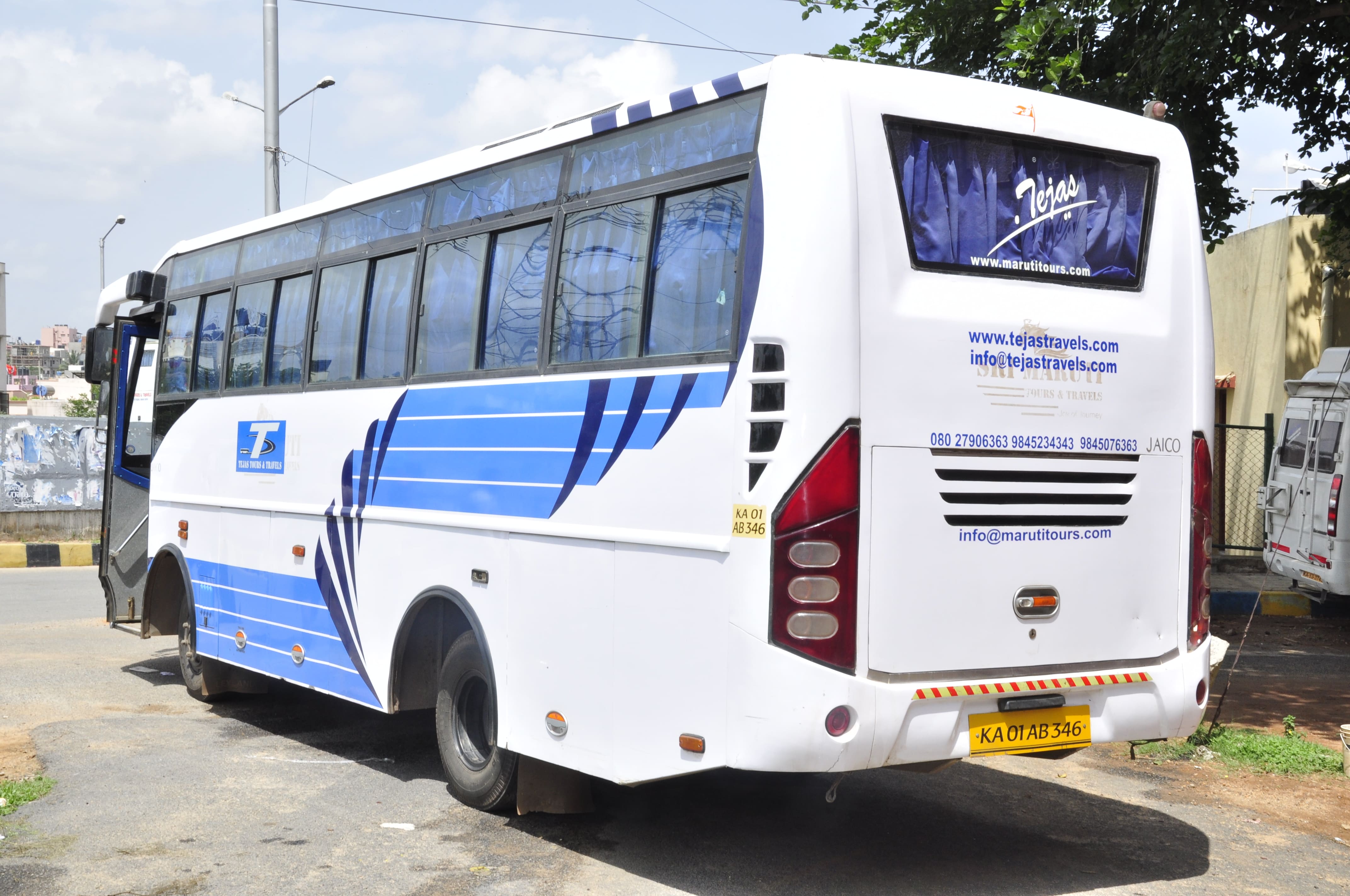 A white bus with blue stripes parked on a street, featuring a sign for Tejas Travels and contact information on the back.