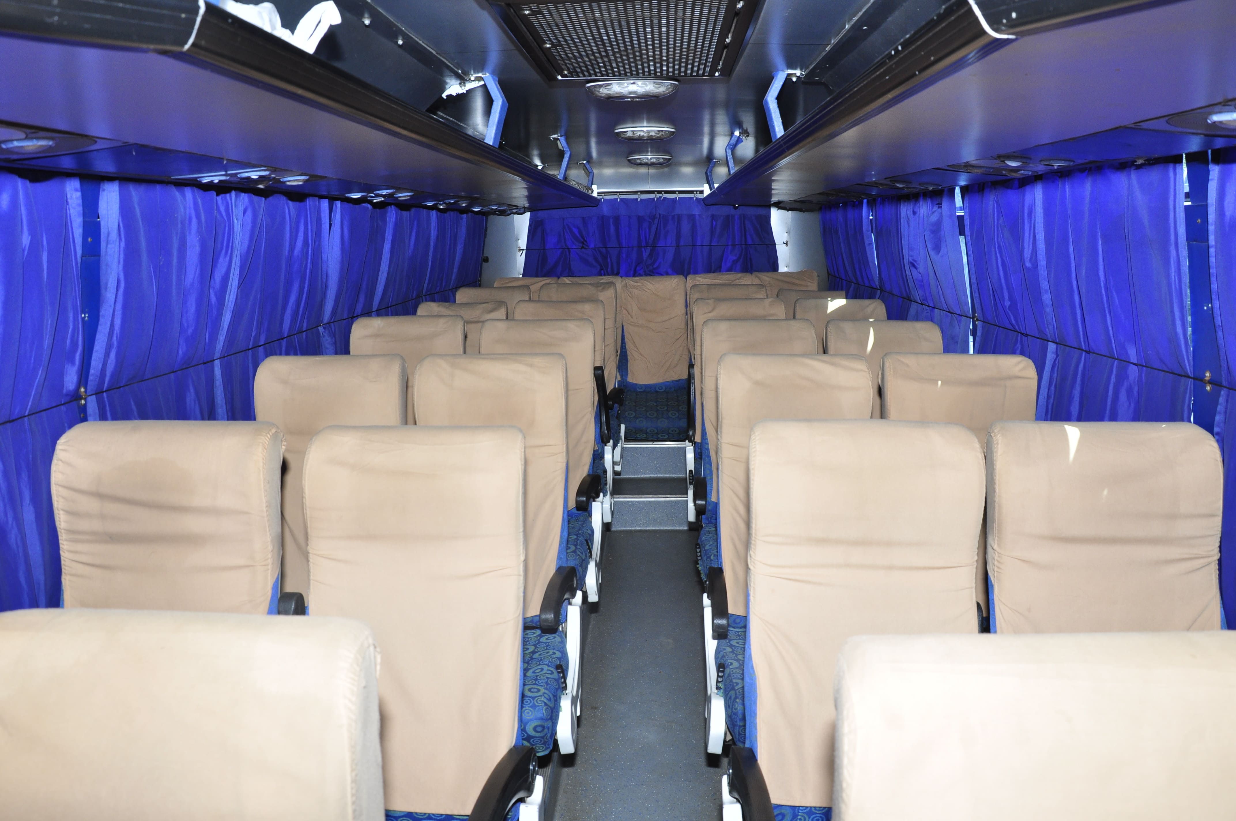 Interior of a bus featuring rows of beige seats and blue curtains, with an empty aisle creating a spacious feel.