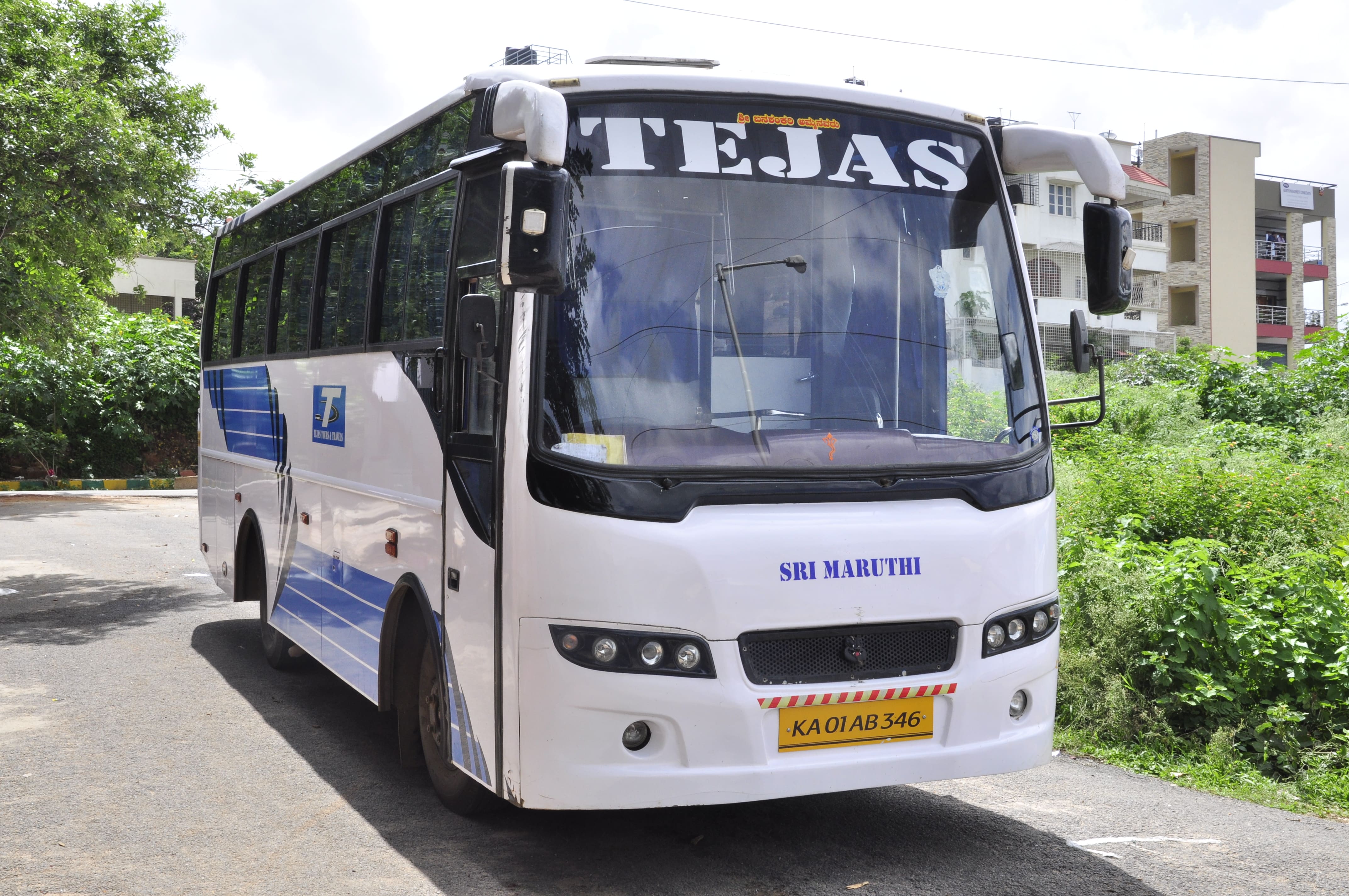White tour bus labeled "TEJAS" parked on a road, surrounded by greenery and residential buildings.
