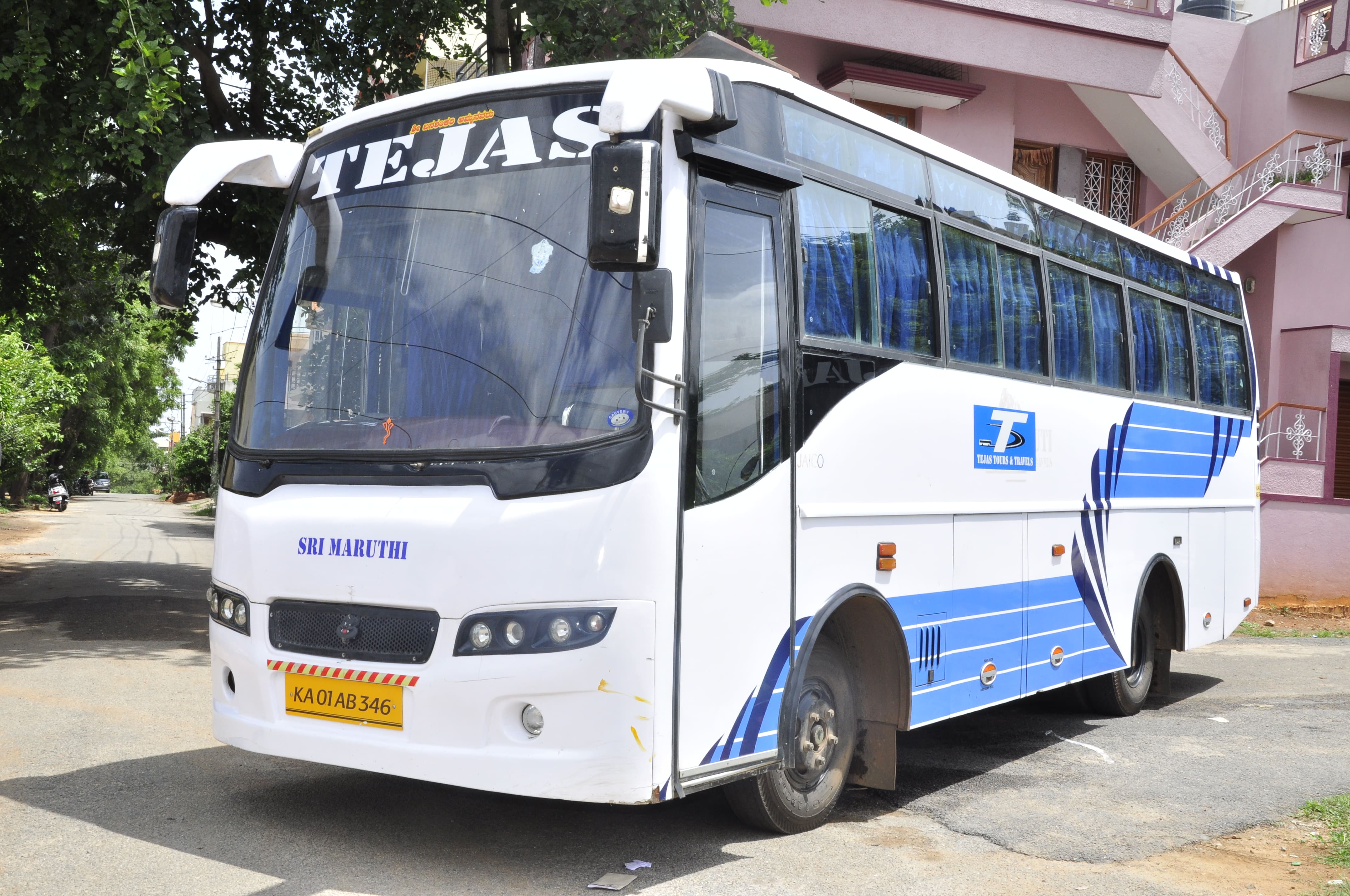 A white and blue bus with "TEJAS" on the front parked on a quiet street lined with trees and buildings.